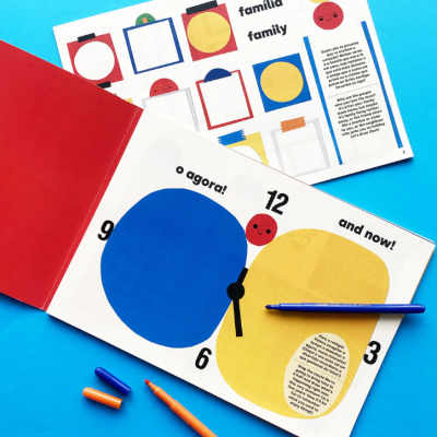 Livro aberto com gráficos coloridos de formas circulares e texto em português e inglês, sobre mesa azul com canetas ao lado.