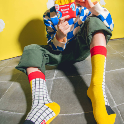 Criança sentada com meias coloridas xadrez, calças verdes e camisola azul e branca segurando embalagem vermelha de batatas fritas.