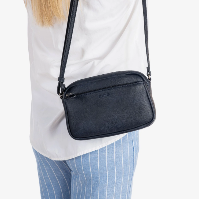 Mala tiracolo preta em material texturizado com fecho de correr usada com camisa branca e calças azuis às riscas