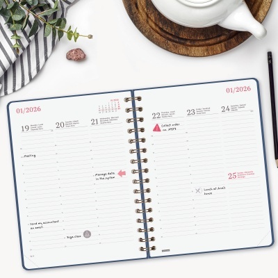 Agenda semanal aberta com anotações, rodeada por chávena, bule e guardanapo às riscas com ramos verdes.