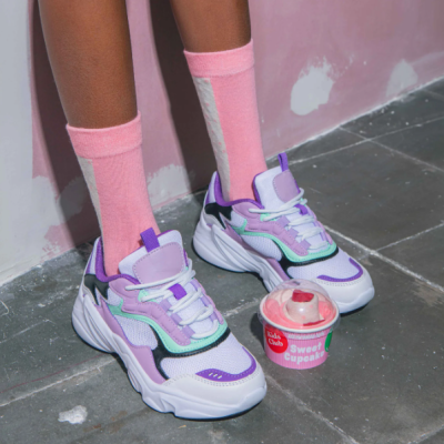 Sapatilhas brancas e lilás com meias rosa junto a recipiente rosa com tampa transparente e texto