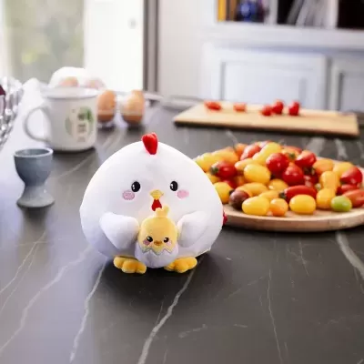 Peluches de galinha e pintainho em mesa preta, com tomates, ovos e chávenas ao fundo