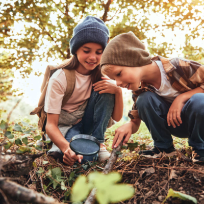 Duas crianças com gorros explorando o solo com lupa numa floresta