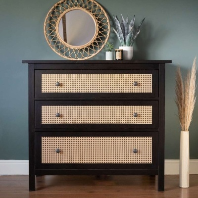 Cômoda preta com gavetas de rattan, espelho redondo de rattan, parede verde, vaso com planta, vaso com planta seca.