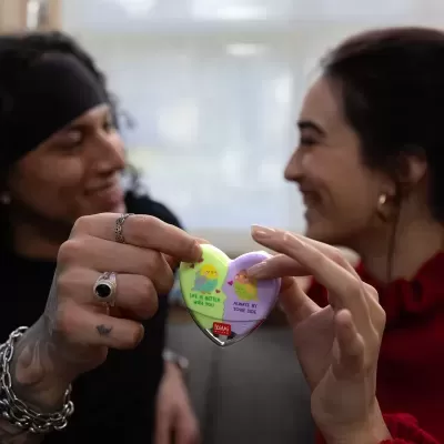 Produto coração verde e lilás com pássaros e texto, segurado por duas pessoas