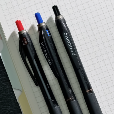 Três canetas Pentonic pretas com tampas coloridas sobre papel quadriculado