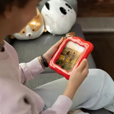 Moldura fotográfica vermelha com ursinho, com foto de casal, numa mão de pessoa sentada num sofá com almofadas de pelúcia