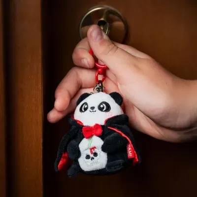 Chaveiro de peluche panda vestido com capa preto e vermelha pendurado em porta de madeira