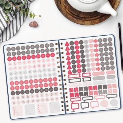 Caderno aberto com adesivos coloridos em rosa, vermelho e cinza sobre uma mesa branca com acessórios.