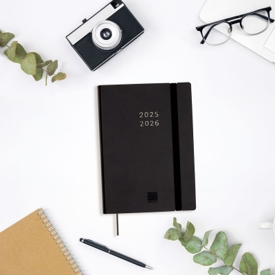 caderno preto com anos 2025 2026 na capa, caneta, folhas verdes, câmera, óculos e laptop sobre mesa branca