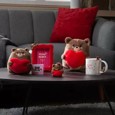 Conjunto de ursos de peluche com corações vermelhos, moldura e caneca sobre mesa cinzenta em frente a sofá cinzento com almofada vermelha.