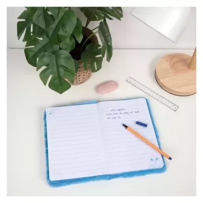 Caderno aberto com capa azul felpuda e caneta sobre mesa branca com planta verde e candeeiro