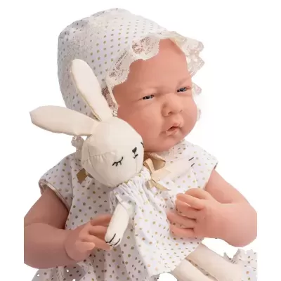 Boneca bebê realista com vestido branco às bolinhas douradas e touca de renda segurando coelho de peluche