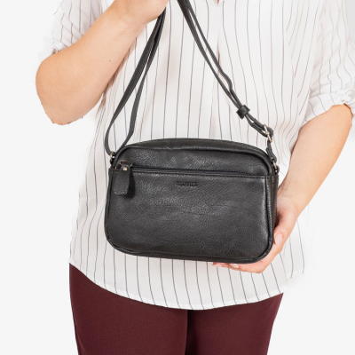 Mala de ombro preta em material semelhante a couro com fecho de correr frontal segurada por pessoa com camisa branca às riscas e calças vinho.
