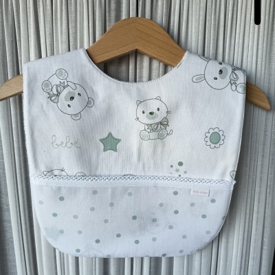 Babete branco com desenhos infantis e padrão de bolinhas verdes pendurado em cabide de madeira