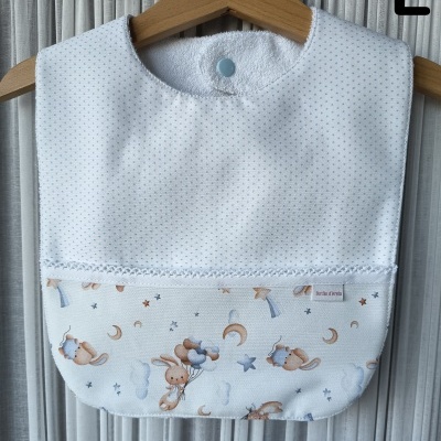Babete branco com pequenos pontos azuis, estampado com coelhinhos, luas e estrelas, pendurado em cabide de madeira.