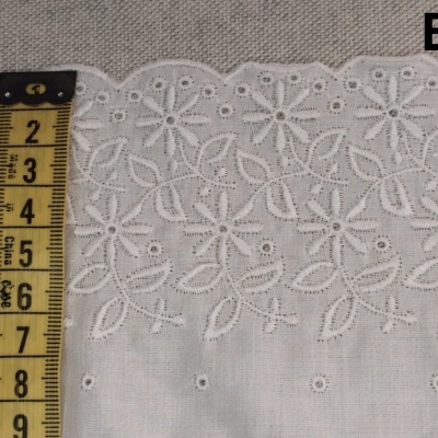 Tecido branco com bordado floral medido por fita métrica amarela, texto B5