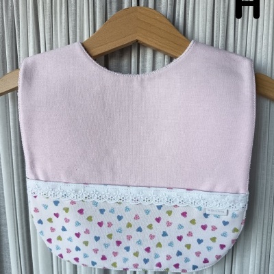 Babete de bebé cor-de-rosa e branco com corações coloridos e renda pendurado num cabide de madeira