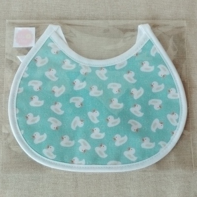 Babete azul claro com padrão de patinhos brancos e borda branca dentro de saco plástico