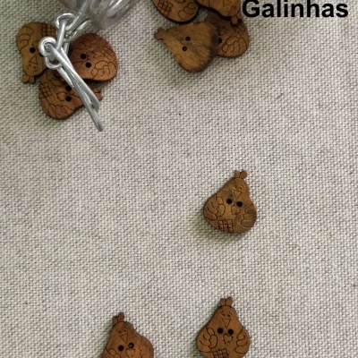 Botões de madeira em forma de galinha com padrões gravados sobre tecido bege e frascos transparentes com texto 'Galinhas'