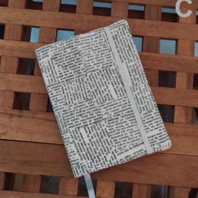 Capa de caderno com padrão de texto preto e branco e elástico cinza sobre superfície de madeira.