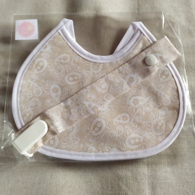 Babete bege com padrão branco, bordas brancas e presilha em saco plástico