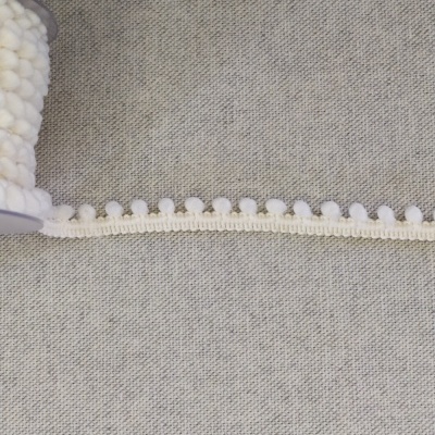 Rolo de fita decorativa creme com pequenos pompons sobre tecido cinza