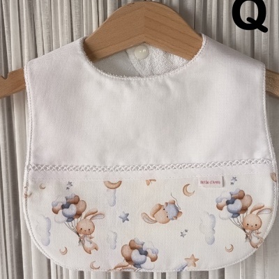 Babete branca com padrão infantil de coelhos e balões em tons pastéis pendurada num cabide de madeira.