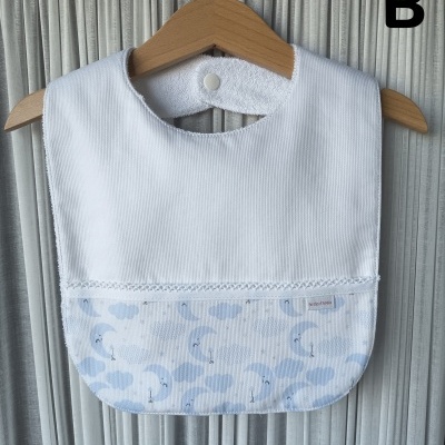 Babete de bebé branco e azul com padrão de nuvens e luas pendurado num cabide de madeira.