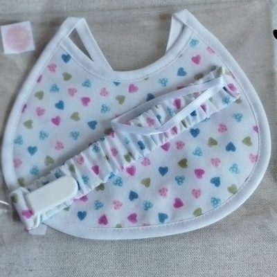 Babete branco com corações coloridos e faixa de cabelo com padrão igual, num saco plástico