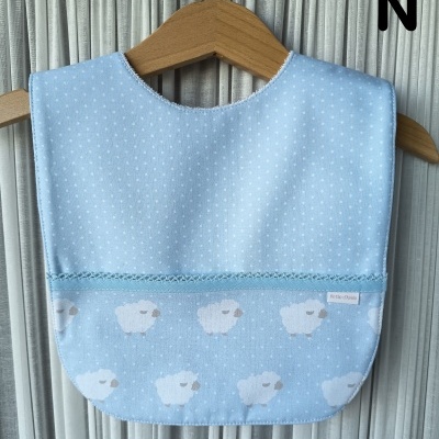 Babete azul claro com bolinhas e ovelhas brancas pendurado num cabide de madeira