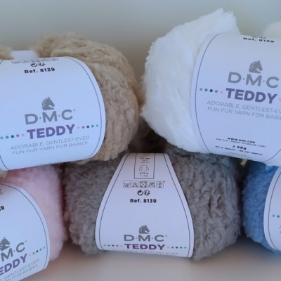 Cinco novelos de lã DMC TEDDY em cores bege, branco, rosa, cinzento e azul com etiquetas brancas.