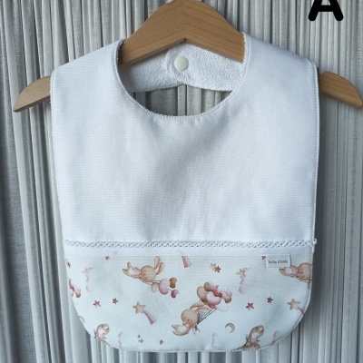 Babete de bebé branco com estampa de coelhinhos e estrelas em tons suaves, pendurado numa cabide de madeira.