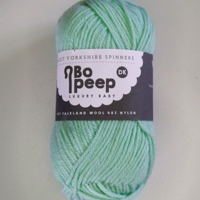 Novelo de lã verde claro com rótulo roxo com texto sobre fundo cinza claro.