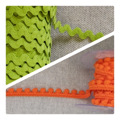 Duas fitas decorativas em rolos, uma verde-limão ondulada e outra laranja com pompons, sobre tecido cinza.