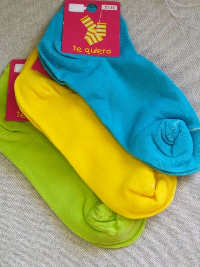 Três pares de meias coloridas azul, amarelo e verde com etiquetas rosas sobre tecido cinza.