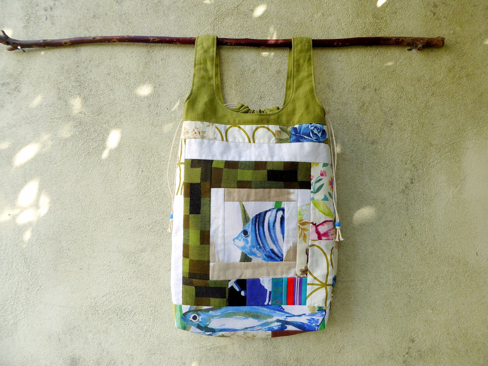 Sacos Com Asas / Saco do Pão Bolsa patchwork com desenhos de peixes e flores, alças verdes pendurada num pau castanho numa parede cinza