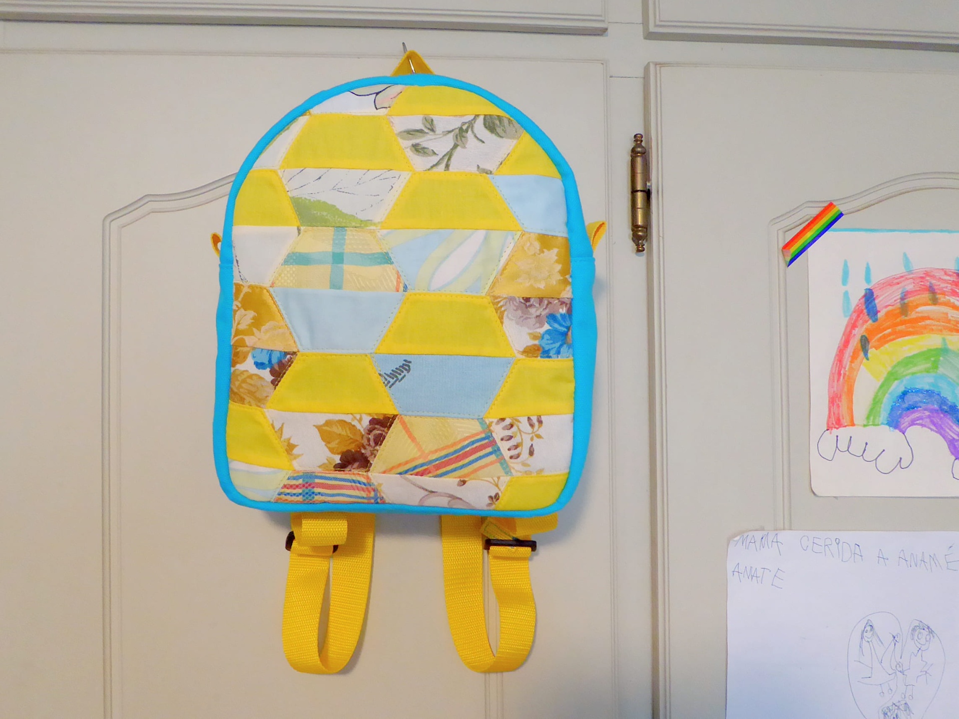 Mochila Pequena Mochila patchwork colorida pendurada numa porta branca com desenhos de arco-íris ao lado.