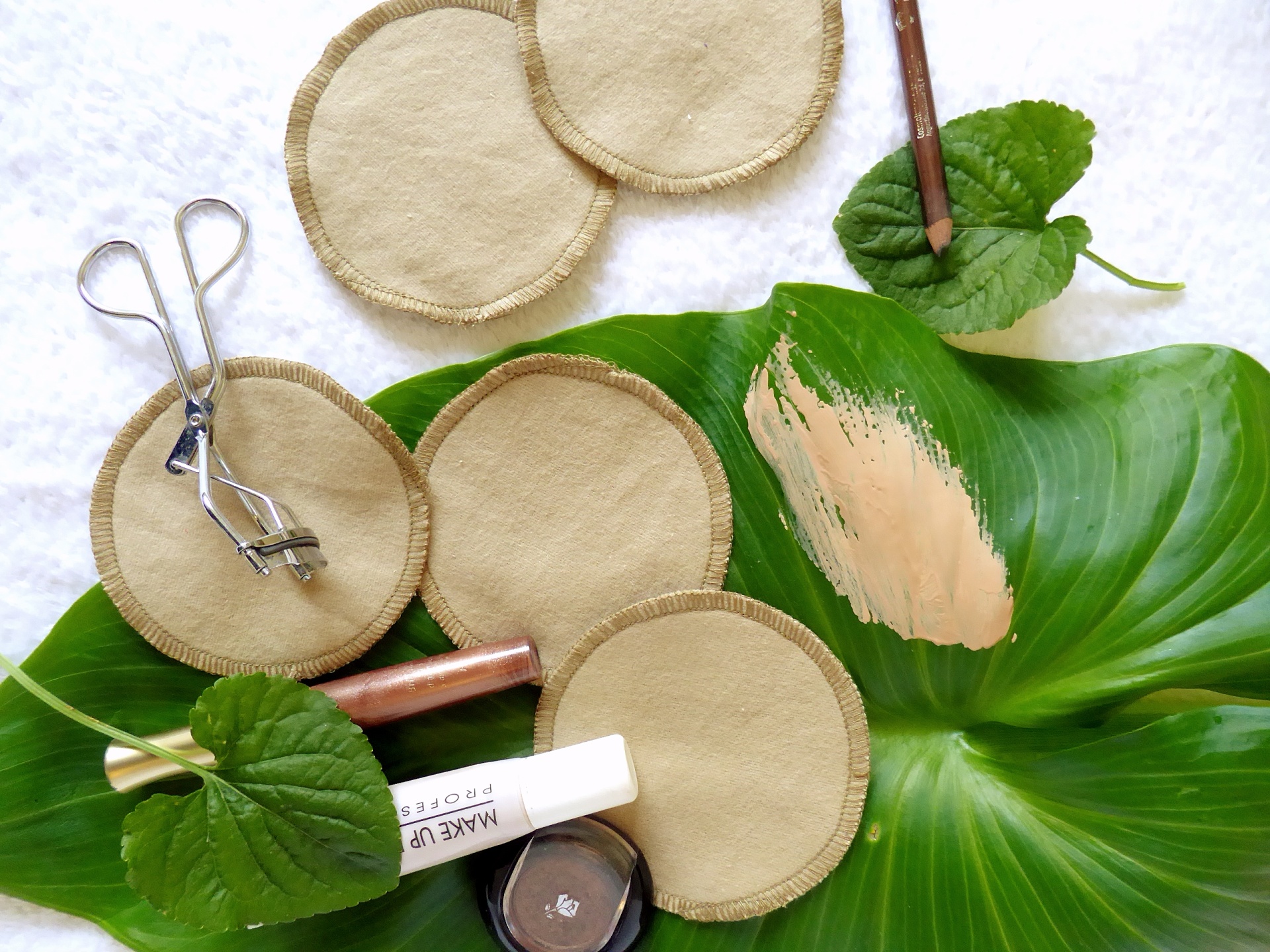 Discos de Limpeza Facial Reutilizáveis Discos desmaquilhantes em tecido, curvador de pestanas e produtos de maquilhagem sobre folhas verdes