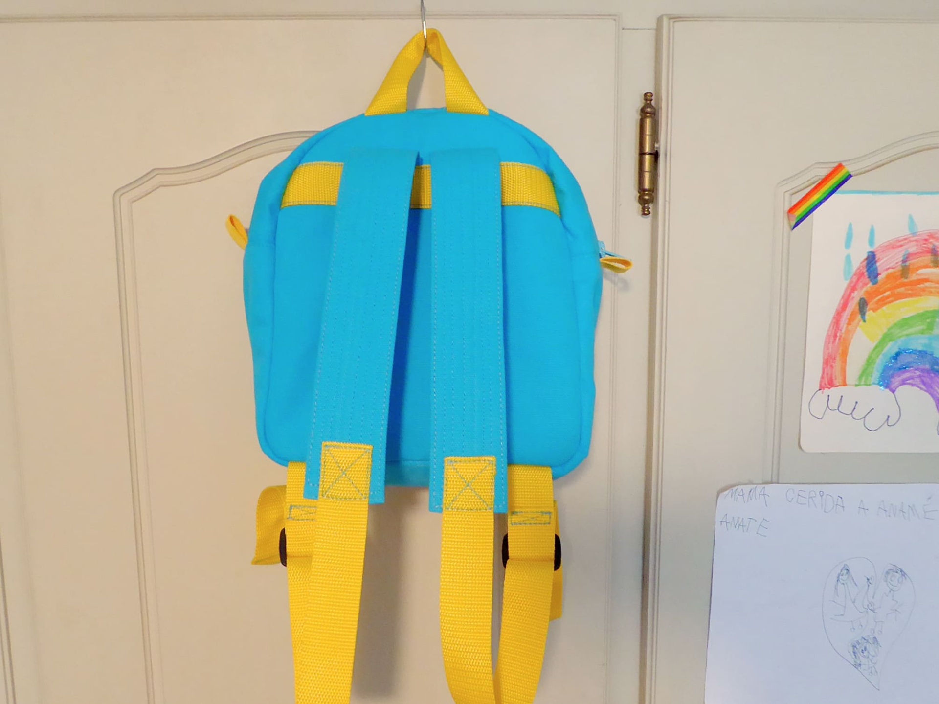 Mochila Pequena Mochila azul com alças amarelas pendurada numa porta branca com desenho infantil colorido ao lado
