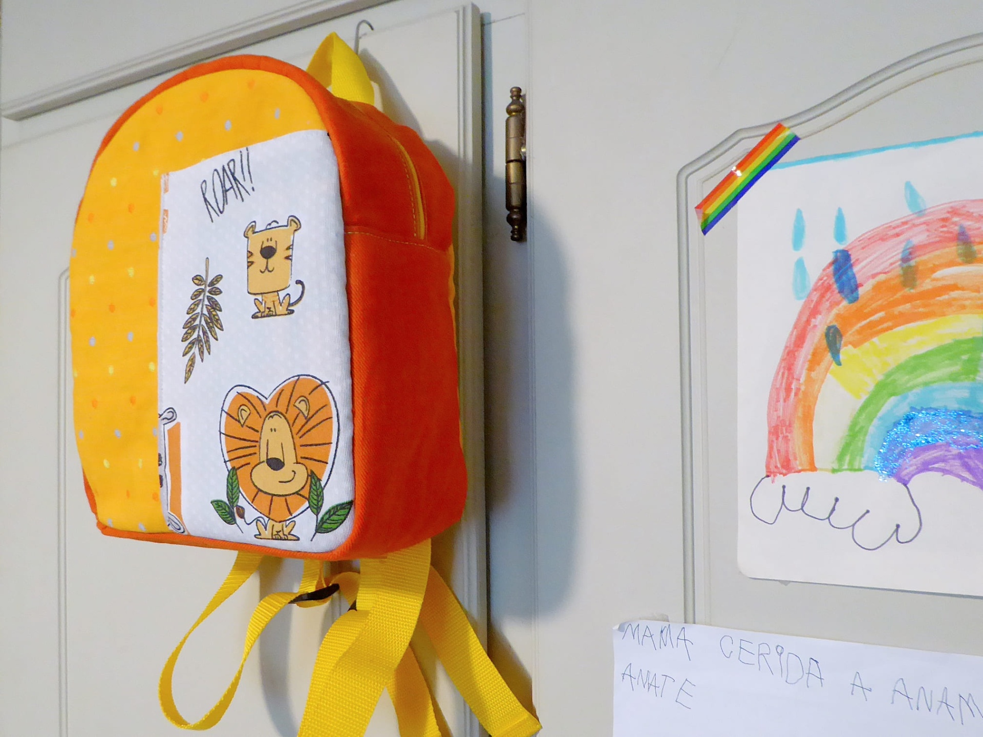Mochila Pequena Mochila infantil colorida com desenhos de leões e o texto ROAR pendurada numa porta em branco próxima de desenho de arco-íris.