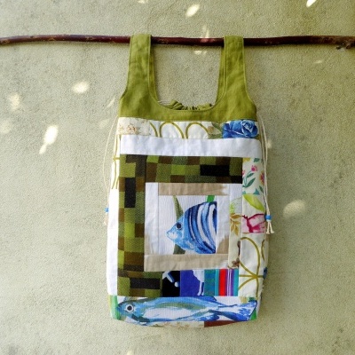 Bolsa patchwork com desenhos de peixes e flores, alças verdes pendurada num pau castanho numa parede cinza
