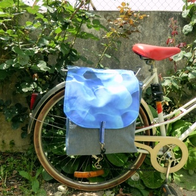 Saco de bicicleta azul e cinza montado numa bicicleta branca com selim vermelho junto a muro e plantas.