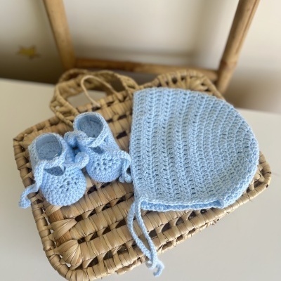 Conjunto de chapéu e sapatinhos de bebé em crochet azul claro sobre cadeira de madeira com assento em palha trançada