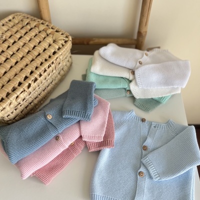 conjunto de casacos de malha de bebé coloridos com botões de madeira sobre superfície branca ao lado de cesta de palha