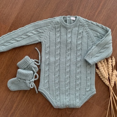 Macacão e botinhas de bebé em malha cinzenta com padrão de tranças sobre superfície de madeira ao lado de espigas de trigo secas