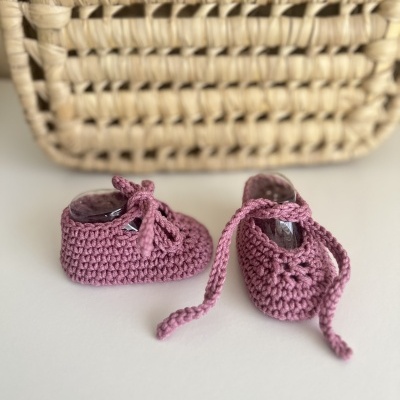 Sapatinhos de bebé em croché cor de rosa com cadarços diante de uma cesta de palha.