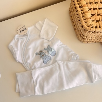 Conjunto para bebé branco com bordados azuis de coelhos e calças, sobre uma mesa clara perto de uma cesta de vime