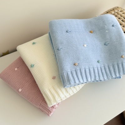 Três mantas de malha rosa claro, creme e azul claro com pompons coloridos sobre superfície branca