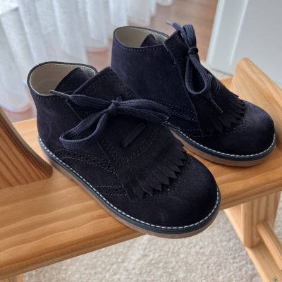 Sapatos infantis pretos em camurça com franjas, atacadores e sola castanha em cadeira de madeira.